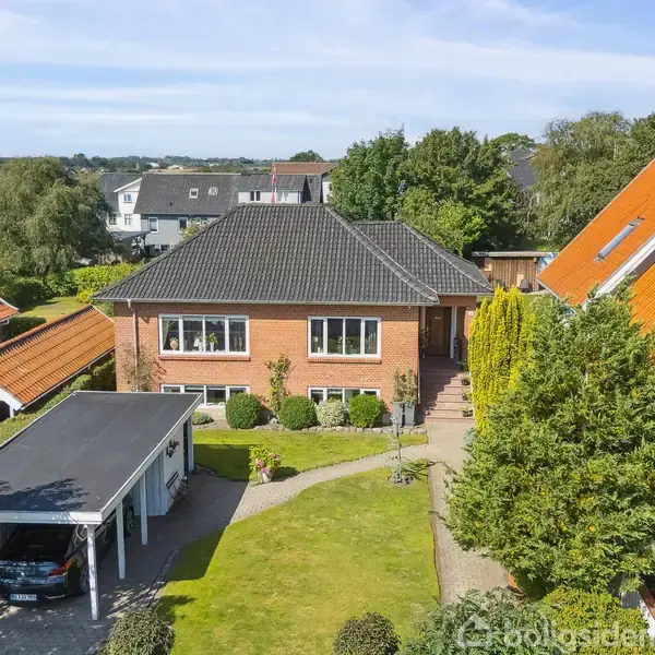 Rødt murstenshus med sort tag i et villakvarter. Foran er en have med græs, buske og en sti. Til venstre er en carport med parkeret bil.