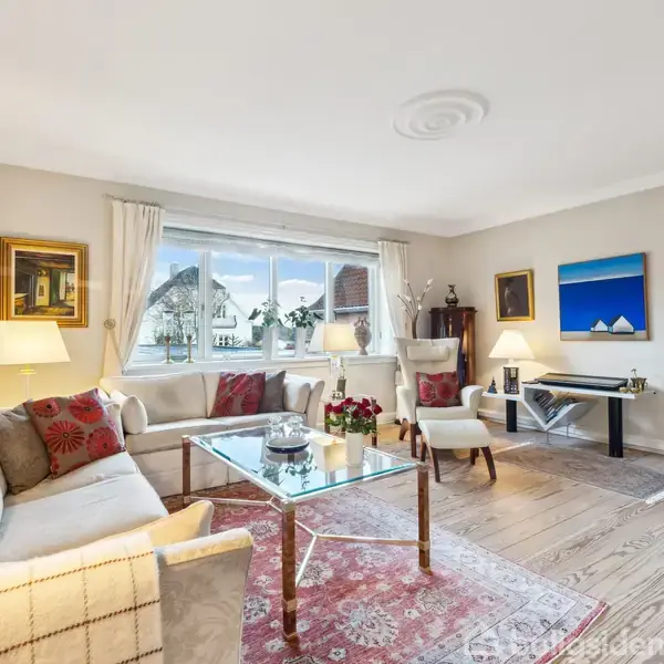 En lys, elegant stue med to cremefarvede sofaer rundt om et glas-sofabord på et tæppe. Et stort vindue slipper naturligt lys ind, og væggene har kunstværker.