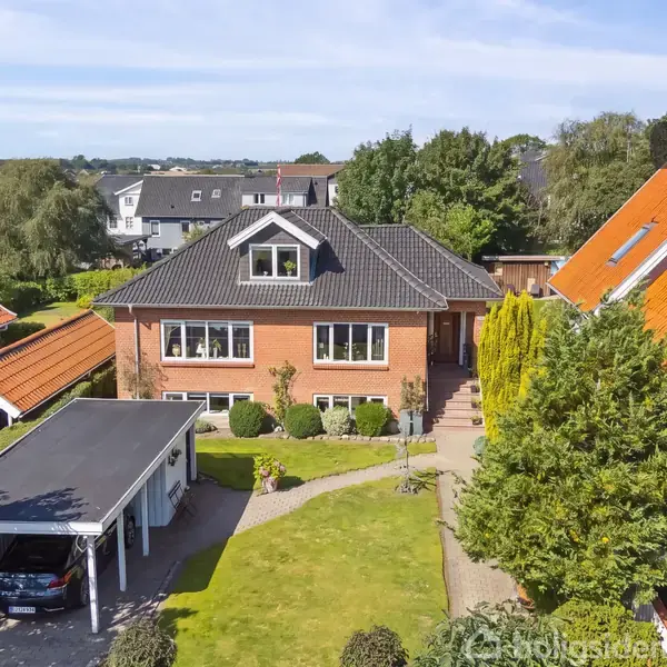 Et murstenshus med sort tag står i en have omgivet af andre huse med orange tage. Foran er en carport med en bil under. Refurt "Visualisering".