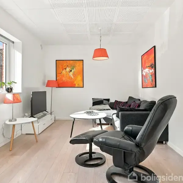 En stue med sort lænestol og sofa. På væggen hænger to farverige malerier. Et stort vindue giver udsigt til en bygning udenfor. Indretningen inkluderer planter og moderne møbler.