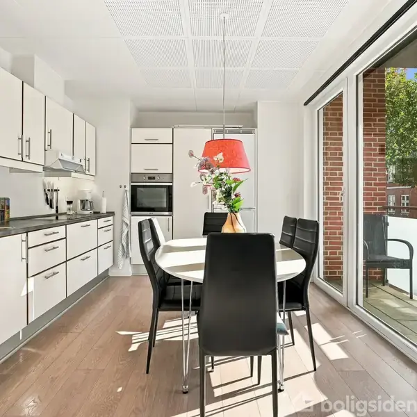 Et moderne køkken-alrum med hvide skabe og sort bordplade. Et rundt spisebord med sorte stole står ved en stor glasdør til en altan. Rød lampe over bordet.