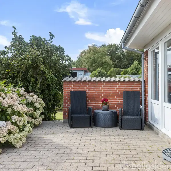 Terrasse med to sorte liggestole ved et rundt bord. Omkranset af rødstensmur og blomstrende buske. I baggrunden ses et grønt område under en blå himmel.