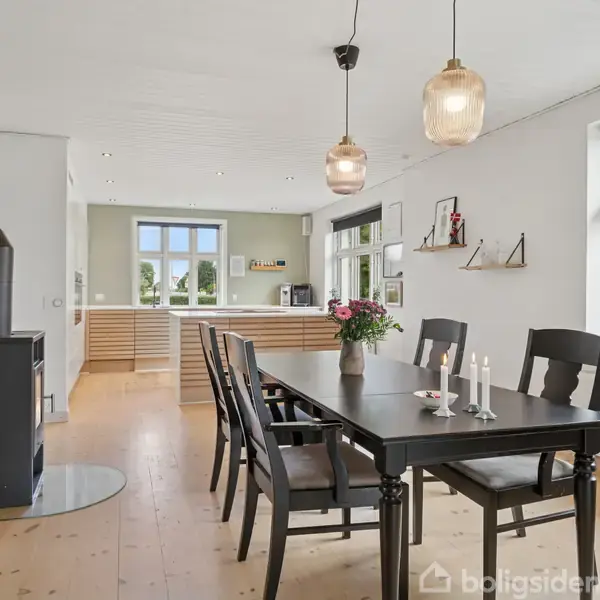 Spisebord med sorte stole står i en lys spisestue. En vase med blomster og stearinlys på bordet. Til venstre er en brændeovn, og i baggrunden ses et åbent køkken.