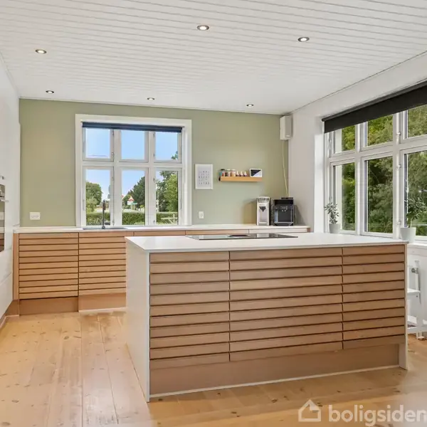 Køkken ø med trælåger står i midten, omgivet af hvide vægge med store vinduer, som slipper naturligt lys ind. Grøn væg i baggrunden med indbygget ovn og kunst på væggen.