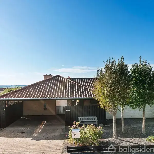 Et hus med rødt tegltag står stille. Det har en indkørsel med småsten og omgivende træer. Huset ligger i et boligområde med blå himmel i baggrunden.