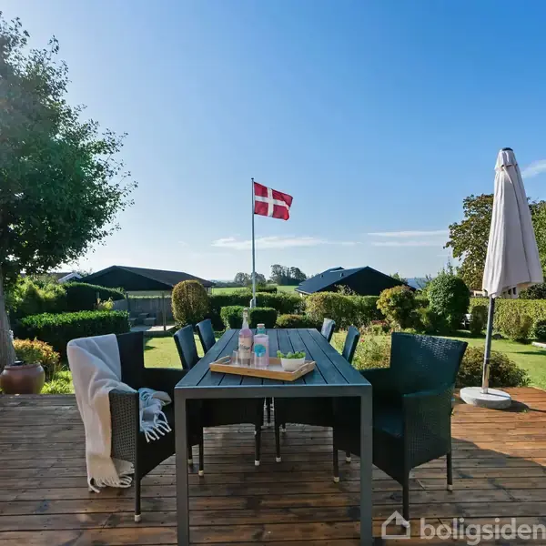 Et udendørs træterrassebord med seks stole, nogle med tæpper, står under en klar blå himmel. I baggrunden vejer et dansk flag. Haven har buske, træer og en parasol.
