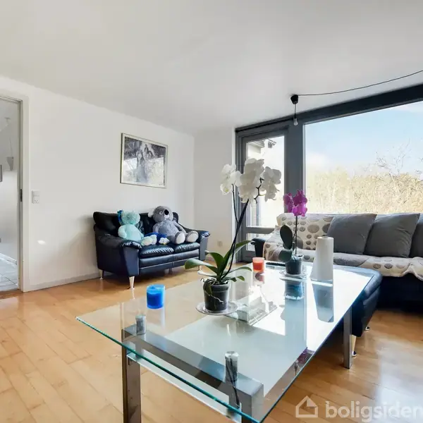 Glasbord med planter stående i stue med træparketgulv. Sofaer placeret ved vinduer med udsigt til natur. Åben dør fører til køkken. Ingen tekst bortset fra "Boligsiden".