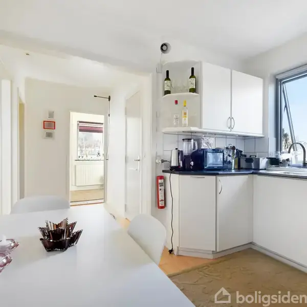 Et moderne køkken med hvide skabe og store vinduer. Der står frugt på et bord i forgrunden, og køkkenapparater som kaffemaskine er placeret på bordpladen.