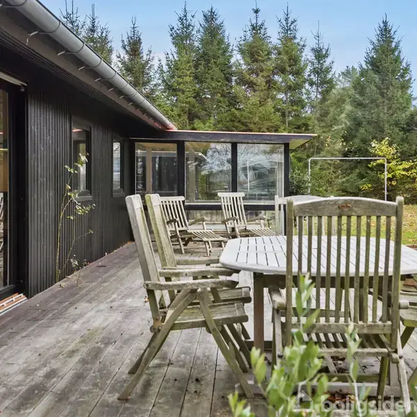 En rund terrassebord med tilhørende stole står på en træterrasse. I baggrunden ses en sortmalet bygning med store vinduer og en have med træer.