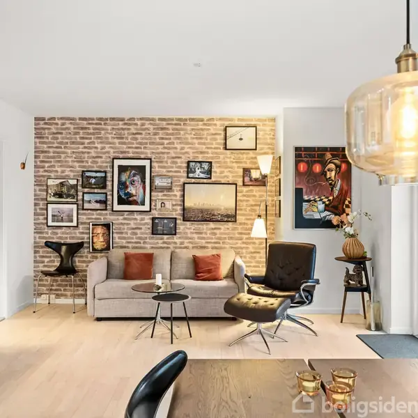 En stue med sofa og lænestol står foran en murstensvæg dekoreret med kunst. En lampe hænger fra loftet, mens franske døre åbner til en have.