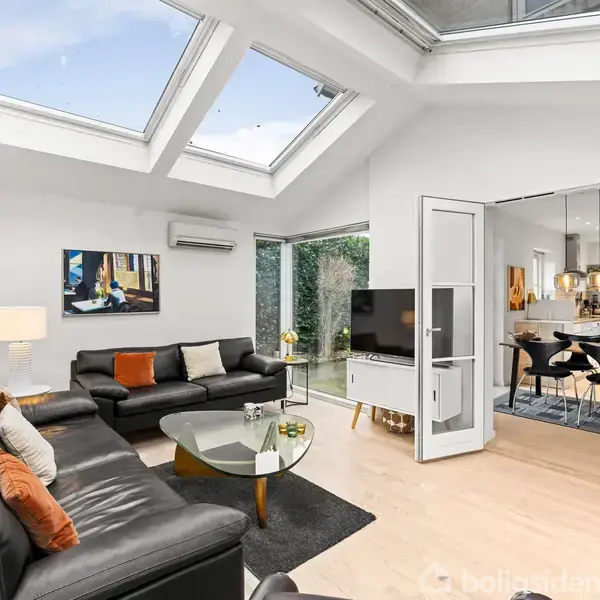 Stue med sort lædersofa, sofabord og kunstværker på væggene. Store ovenlysvinduer lader dagslys strømme ind. Spisestue ses i baggrunden gennem franske døre.