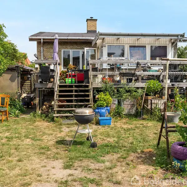 En baghave med en græsplæne, der fører op til en terrasse. På terrassen står flere planter og havemøbler. En grill står på plænen foran en trappe.