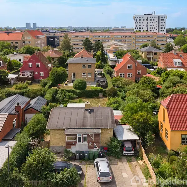 Flere parcelhuse med farverige tage står i en tæt række. Grønne haver omkranser husene, mens en stor lejlighedsbygning ses i baggrunden, indrammet af en blå himmel.