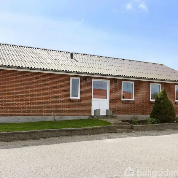 En rød murstensbygning med hvidt tag står stille ved en vejkant. Foran huset er velplejede buske, og græsplæner omgiver bygningen.