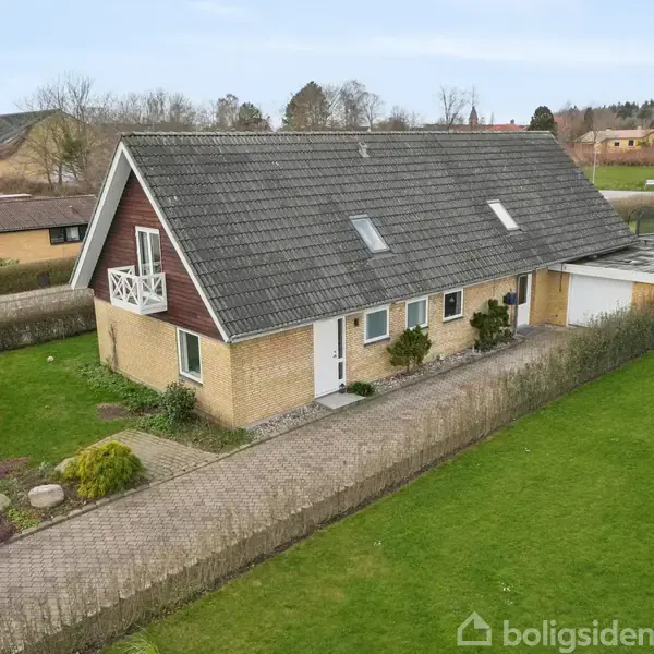 En murstensvilla med gråt tag har en lille altan og en tilknyttet garage. Villaen er beliggende i en forstad med grønne haver og stier omkring.