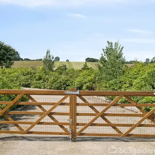 Trælåge står lukket, flankeret af træstakitter, med udsigt til grønne buske og en åben mark i baggrunden under en blå himmel.