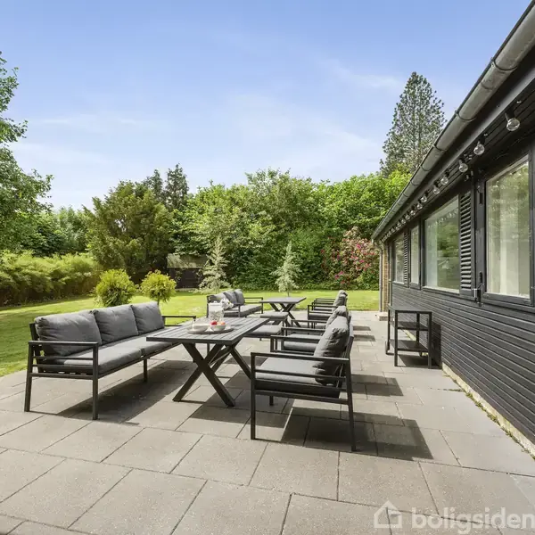 Terrasse med sorte havemøbler står opstillet. Møblerne har grå hynder og står foran et sortmalet hus med vinduer. Omgivet af en frodig have med grønne træer og buske.