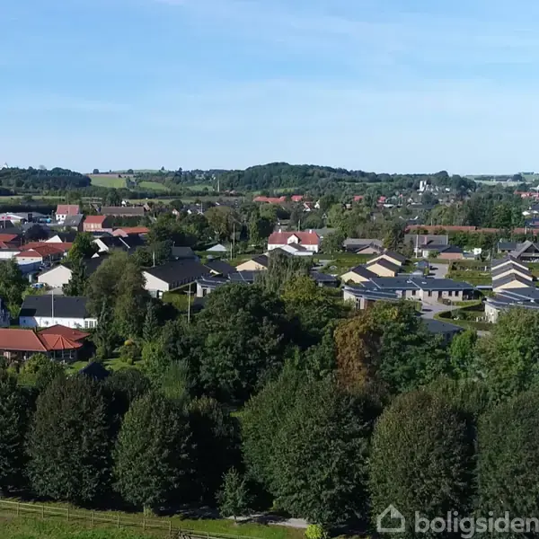 Huse ligger i en tætbebygget forstad, omgivet af frodige træer og grønne marker under en klar blå himmel. Bakkede landskaber ses i horisonten.
