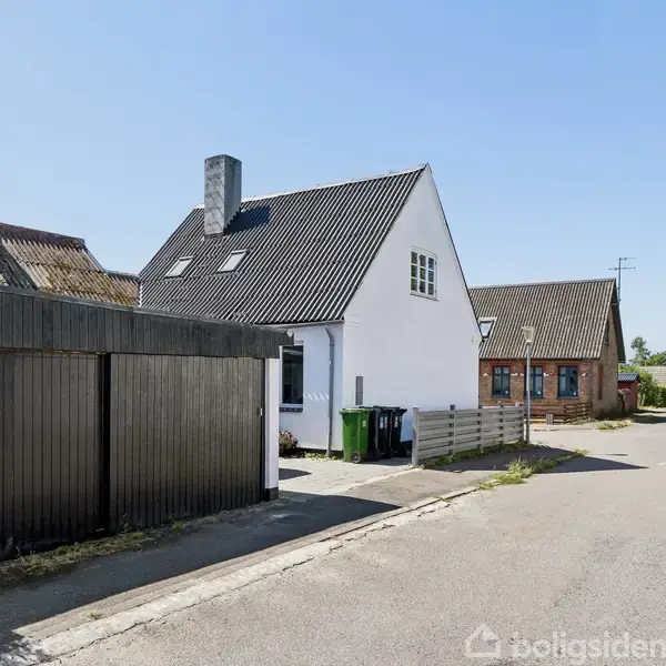Et hvidt hus med skråt tag står på en stille gade. To affaldsbeholdere er placeret ved siden af huset. Omgivet af andre huse og grønne træer i baggrunden.
