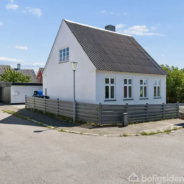 Hvidt hus står stille på et hjørne, omgivet af hegn og en trailer på en rolig vej. Sollys oplyser området, med nogle træer og flere huse i baggrunden.