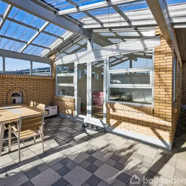 Terrasse med bord og stole står under et gennemsigtigt tag. Murstensvægge omgiver området, og solen skinner ind. Døre fører ind til huset fra terrassen.