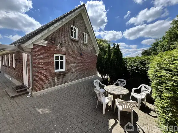 Et rødt murstenshus står på en flisebelagt terrasse. Fire hvide plastikstole omgiver et rundt bord, omgivet af grønne buske og træer under en skyet blå himmel.