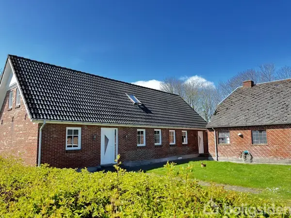 Et rødt murstenshus med sort tegltag står i en grøn have. En anden mindre bygning ses til højre. Himlen er blå med spredte hvide skyer.