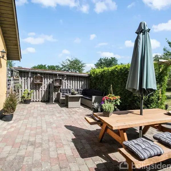 Et træbord med puder og en parasol står på en flisebelagt terrasse. Til venstre er et gult hus med døre, og i baggrunden er en have med planter og møbler.