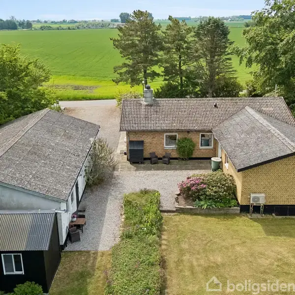 Et hus i gule mursten med et gråt tag står på en grusbelagt grund omgivet af græsplæne og træer. I baggrunden ses en stor, grøn mark.