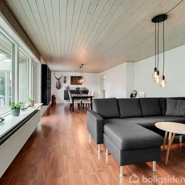 Sort sofa står i en stue ved siden af et lille rundt bord. Der hænger moderne lamper over bordet. Store vinduer viser en have udenfor. Gulvet er parket.