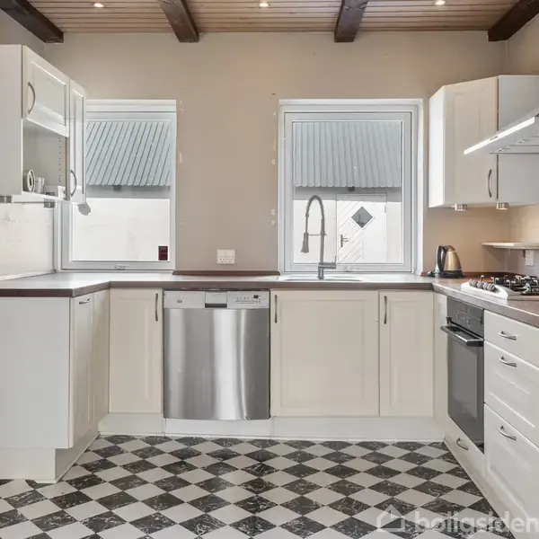 Køkken med hvide skabe og en opvaskemaskine i stål. Der er en gaskomfur og en kedel på bordet. Skakternet gulv og loft med synlige bjælker.