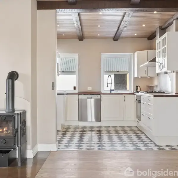 Brændeovn opvarmer rum ved siden af åbent, moderne køkken med hvidt inventar. Køkkenet har flisegulv og ovenlys, mens brændeovnen står på trægulv.