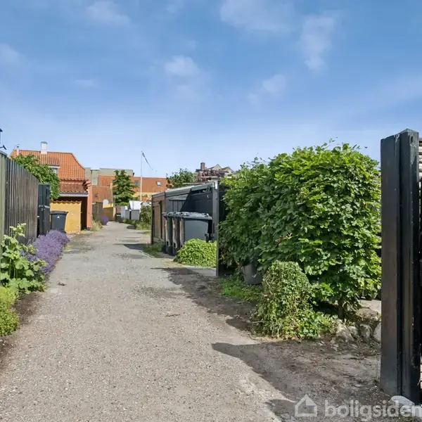 En smal grussti strækker sig mellem to hække med nummer 38-skiltet til højre. I baggrunden ses røde tage og blå himmel.