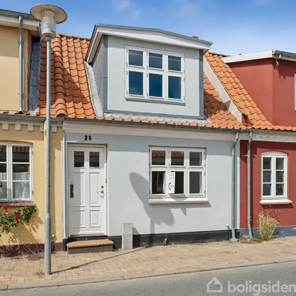 Et rækkehus med et gråt facade og rødt tegltag står i en række farverige huse. Nummer 36 ses ved hoveddøren. En gadelampe og blomstrende busk er i forgrunden.
