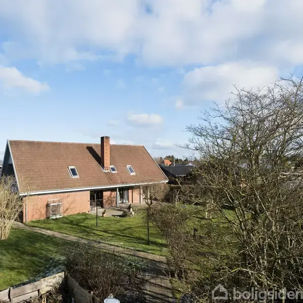 Huset er en enkeltstående bygning med rødt tegltag og flere tagvinduer. Det står i en rummelig have omkranset af træer og buske under en blå himmel med spredte skyer.