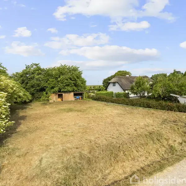 En åben græsplæne ligger foran et lille skur, omgivet af buske. I baggrunden ses stråtækte huse langs en vej. Skyfri himmel med få skyer danner baggrunden.