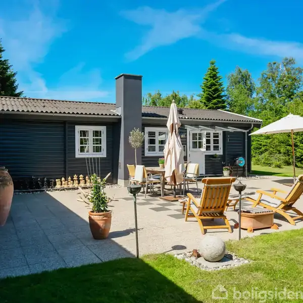 Et sommerhus omgivet af høje træer og grøn bevoksning står på en træterrasse. Flere små bygninger er synlige i baggrunden i en frodig skovklædt have.