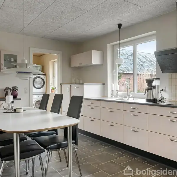 Køkkenbord med seks sorte stole er centralt placeret. Køkkenskabe og apparater, herunder opvaskemaskine og komfur, omgiver bordet. Store vinduer giver lysindfald i det moderne køkkenområde.