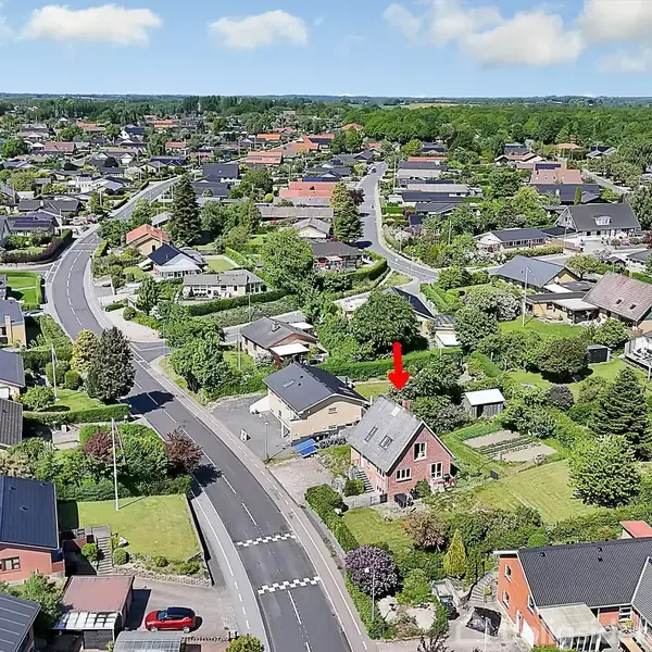 En rød murstensvilla med sort tag ligger i et boligområde. Huset er markeret med en rød pil. Omkringliggende haver og veje er synlige i sommerligt vejr.