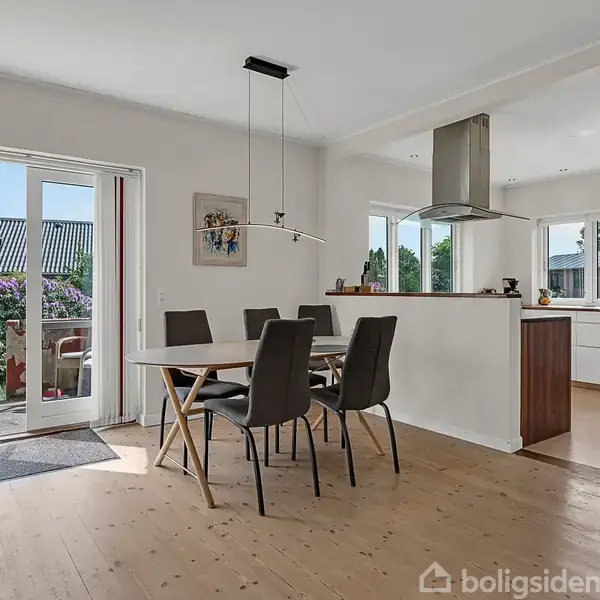 Spisebord med seks stole står centralt i et lyst køkken-alrum. En åben dør leder ud til en terrasse med grønne planter. Køkkenet har moderne hvide skabe og en stor emhætte.