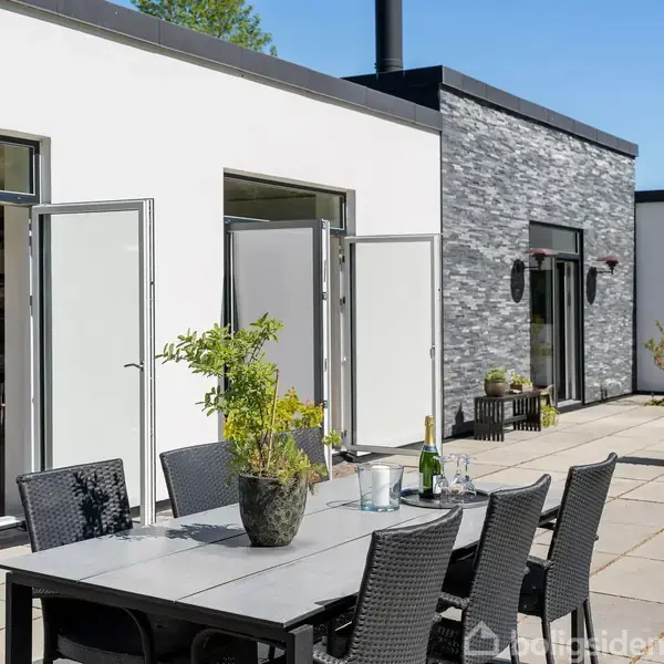 Et udendørs spisebord med stole står på en terrasse ved siden af et moderne hus med åbne døre. Der er grønne planter og sollys omgivende miljøet.