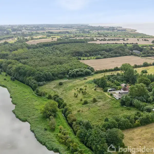 Landområde med marker og skov, der strækker sig langs en kystlinje. En gård omgivet af træer er synlig blandt de grønne områder.