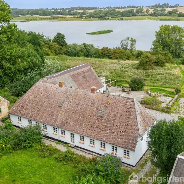 En hvid bygning med stråtækt tag er omgivet af frodig vegetation. Den ligger ved en sø med grønt landskab og træer i baggrunden.