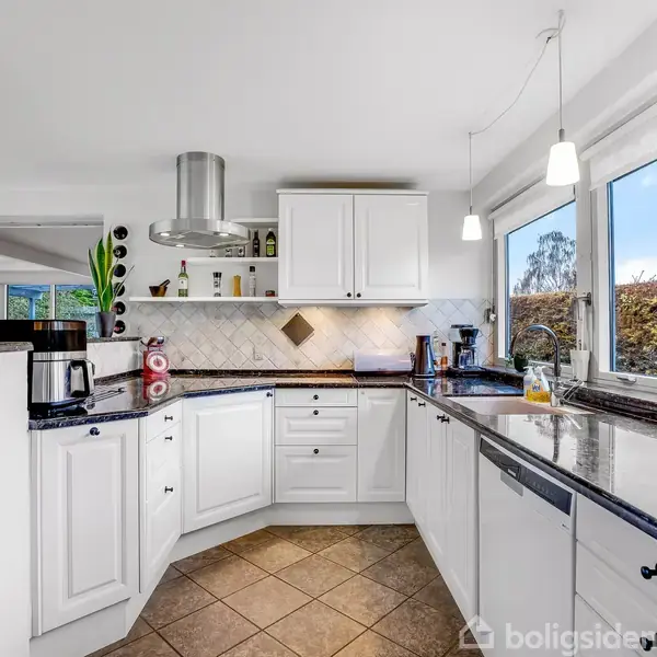 Et rummeligt hvidt køkken med sorte bordplader, kaffemaskine og komfur. Et vindue mod højre slipper naturligt lys ind, oplyser rummet og afslører et udenfor liggende hegn og buske.