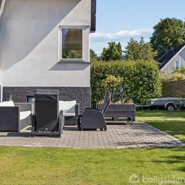 En terrasse med havemøbler står på fliser ved et hvidt hus. I baggrunden ses en græsplæne, en hæk og en trailer. Der er en klar himmel over området.