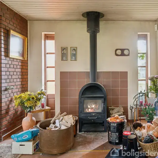Brændeovn står varm i hjørne af rum med rød murstensvæg, omgivet af træhylder fyldt med nipsgenstande og bøger. Ved siden af er der stabler af brænde og en potteplante.