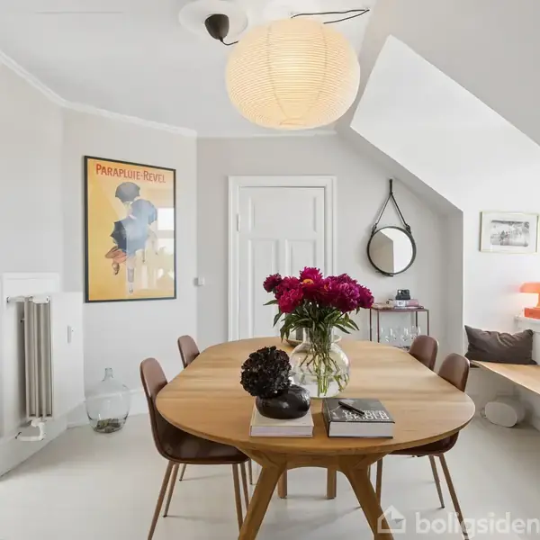 Et rundt træbord med blomster i vasen står i et lyst rum. En plakat ved siden af døren viser "Parapluie-Revel". Der er en hyggekrog med orange lampe i baggrunden.