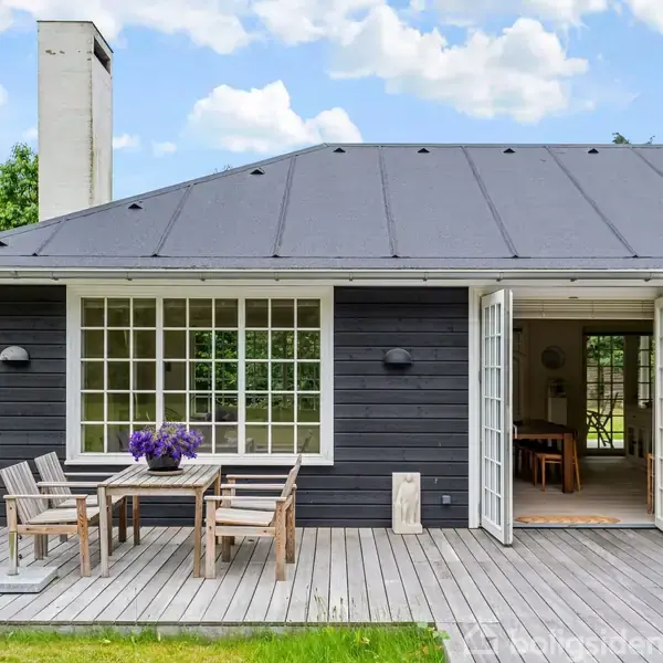 Sort træhus med åbne dobbeltdøre fører ud til en træterrasse med et bord og stole. Blomster står på bordet. Græsplæne og træer omgiver området under en skyet, blå himmel.