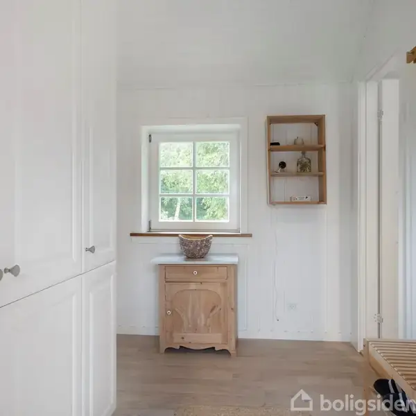 Et skab står stillet op ad en hvid væg. En mindre trækommode er placeret under et vindue med udsigt til grønne træer. Environmentet er lyst med træhylder til højre.