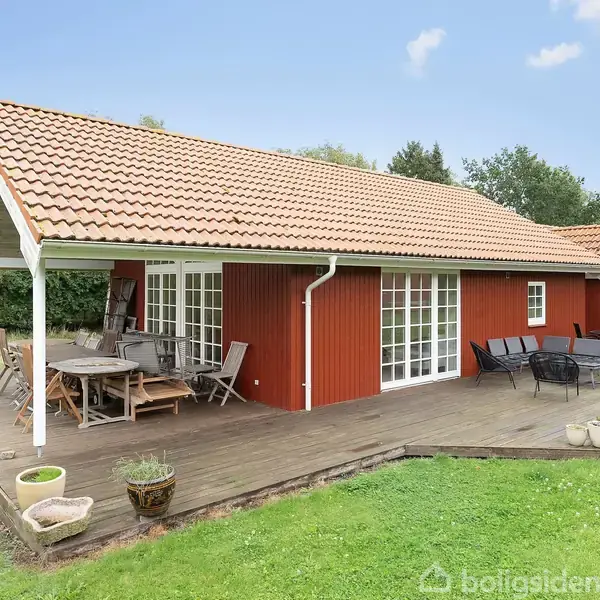 Et rødt træhus med terracottatag sidder på en træterrasse. Huset er omgivet af grøn græsplæne og træer, med havemøbler på terrassen under en overdækket sektion.